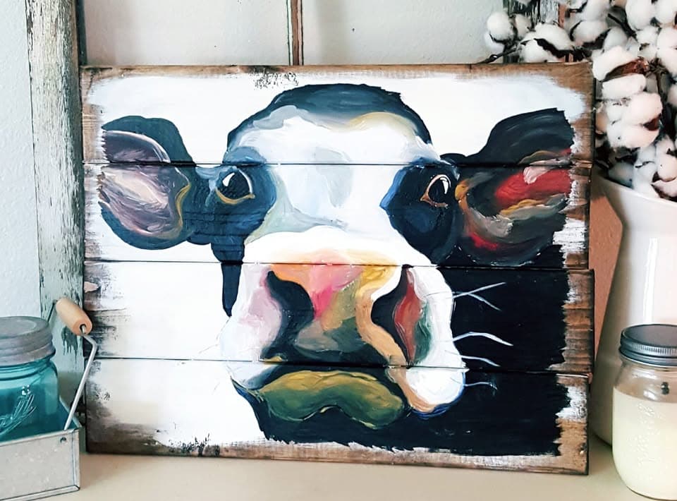 Painting of a black-and-white Holstein cow face on rustic pallet-wood boards, staged with blue mason jars and cotton stems.