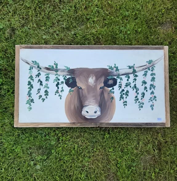 Framed painting of a longhorn bull with a eucalyptus and pink floral garland draped across its horns, photographed on grass.