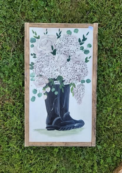 Framed painting of black Hunter rain boots overflowing with white hydrangeas, roses and eucalyptus sprigs, on grass.