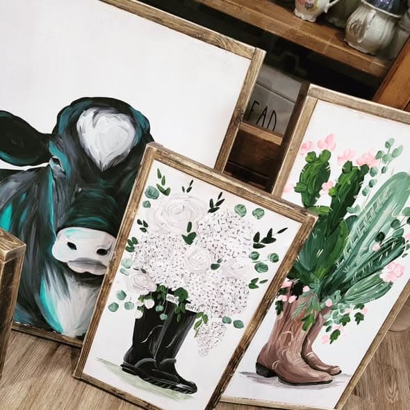 Several framed paintings displayed together: a teal-and-black Holstein cow, Hunter boots with white flowers, and cowboy boots with a pink cactus.
