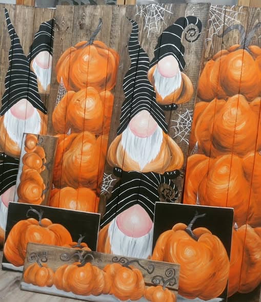 Multiple Halloween porch signs showing gnomes with pumpkins and spider webs alongside standalone orange pumpkin paintings on dark weathered wood.