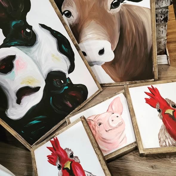 Top-down flat lay of four framed farm animal paintings: a teal Holstein cow, a brown cow, a pink pig and a red rooster.