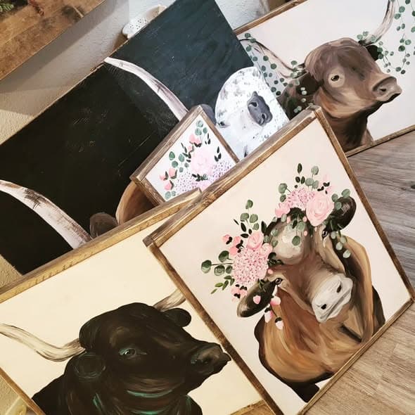 Several framed Highland cattle and longhorn paintings stacked on wood surfaces, including one with a pink floral crown.